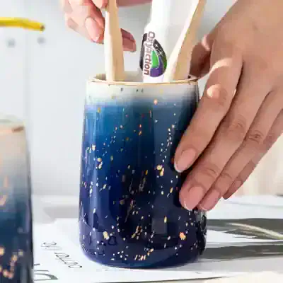 Ceramic mug with gold splatter design and handle, Monalisa store logo visible, artist hand placing wooden sticks inside, modern arts and crafts style, perfect for home decor or gifts, quality ceramic material, unique artistic pattern, vibrant blue color with gold accents, ideal for collection or display at Monalisa Medical.