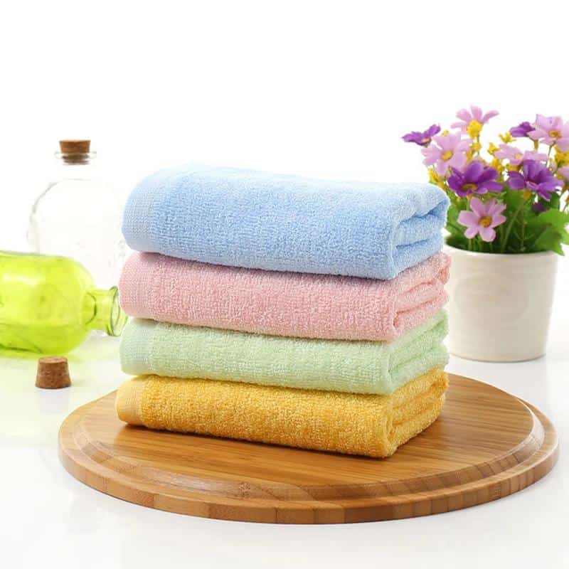 Soft pastel-colored towels stacked on a wooden tray with a potted purple flower and glass bottles in the background, perfect for spa, skincare, or beauty treatments at Monalisa Store.