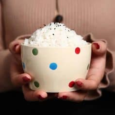Creamy popcorn in a colorful polka dot bowl, held by a person with red nail polish; cozy indoor setting.