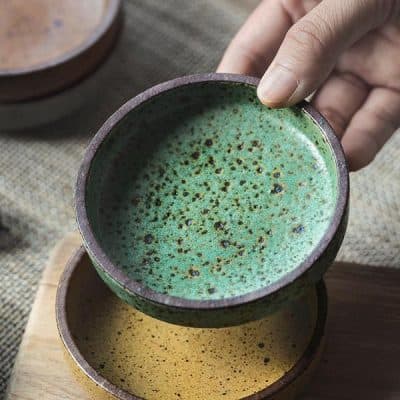 Hand holding a green ceramic bowl with textured glaze, on wooden table, showcasing artisanal pottery.