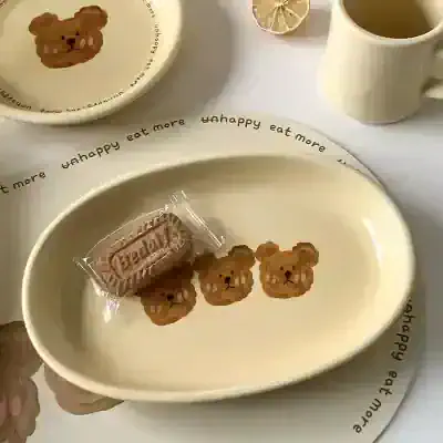 Bear-themed ceramic plate with bear-shaped cookies and a wrapped biscuit, along with a cup of tea and lemon slices on a white table.