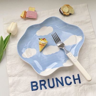 Miniature crepe dessert on a sky-themed plate with a fork, surrounded by small food items and a white tulip, over a white cloth with