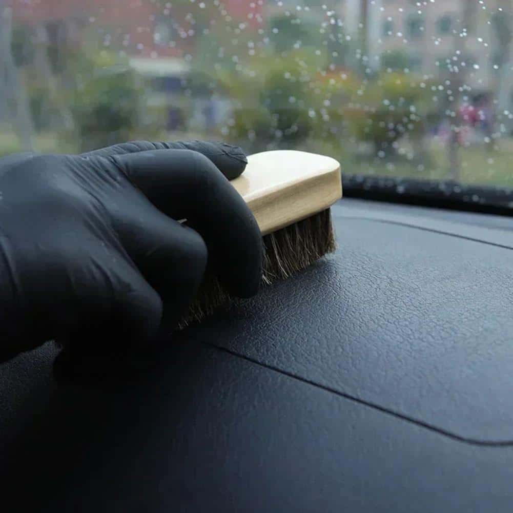 Hand wearing black glove cleaning car dashboard with brush in a rainy environment, showing car interior maintenance.