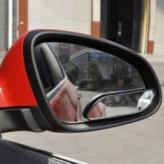 Red car side mirror with integrated blind spot mirror, reflecting parked vehicles and street scene.