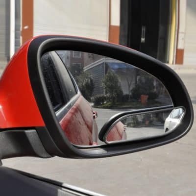 Red car side mirror with integrated blind spot mirror, reflecting parked vehicles and street scene.