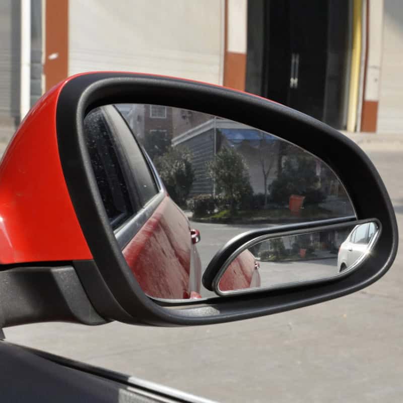 Red car side mirror with integrated blind spot mirror, reflecting parked vehicles and street scene.