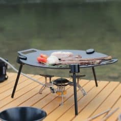Grill with meat and vegetables on a portable outdoor stove set on a wooden table, perfect for outdoor cooking and dining.