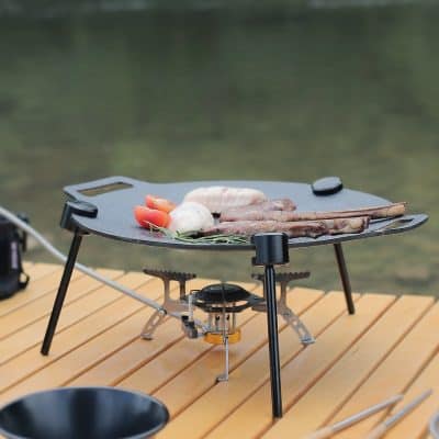 Grill with meat and vegetables on a portable outdoor stove set on a wooden table, perfect for outdoor cooking and dining.