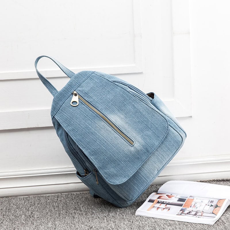 Light blue denim backpack with multiple zippered compartments, ideal for school or travel, prominently displayed on a white wall background.