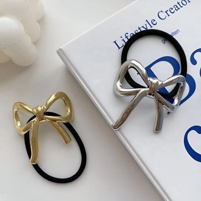 Elegant metallic bow hair accessories on black elastic hair ties, perfect for women’s fashion and styling, featured near a lifestyle magazine and decorative beads.