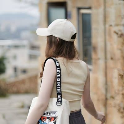 Female wearing a white baseball cap and chic sleeveless top, carrying a stylish tote bag with animated graphic, standing outdoors on a city street with a beige brick building in the background.