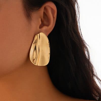 Gold statement earring with irregular shape, worn by a woman with dark hair, close-up fashion jewelry image for luxury accessories and jewelry collections.