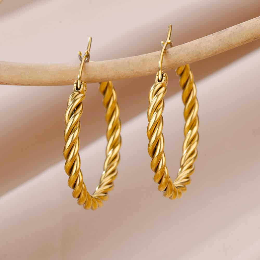 Shiny gold twisted hoop earrings hanging on a beige wooden display. Elegant jewelry for women, perfect for accessorizing any outfit, featuring high-quality craftsmanship and timeless design from Monalisa Store.