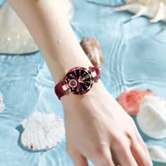 Luxurious women's wristwatch with a deep pink and black gemstone dial, elegant gold accents, and a velvet strap, showcased on a model's wrist against a water and seashells backdrop for stylish fashion appeal.