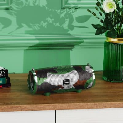 Wireless portable Bluetooth speaker with camouflage design, placed on wooden surface with green vase and flowers in background.