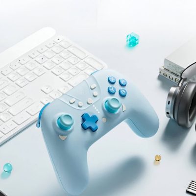 Wireless gaming controller with white and blue design on a clean white desk, beside a keyboard and headphones, emphasizing modern gaming and tech accessories.