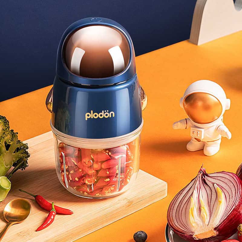 Plodon mini food chopper with astronaut toy, colorful vegetables and chili peppers on orange background, compact kitchen appliance, ideal for quick food prep, safe for kids, modern design.