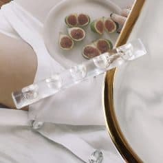 Sculptural clear acrylic eye dropper on a white tray with fresh figs, elegant gold-rimmed mirror, and soft fabrics, perfect for luxury beauty and eye care product presentation at Monalisa Store.