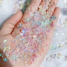 Glittering iridescent bow-shaped nail art embellishments on a hand, acrylic decorations for nails, holographic nail art accessories, Monalisa Medical store quality nail decor, colorful and shiny nail craft supplies.