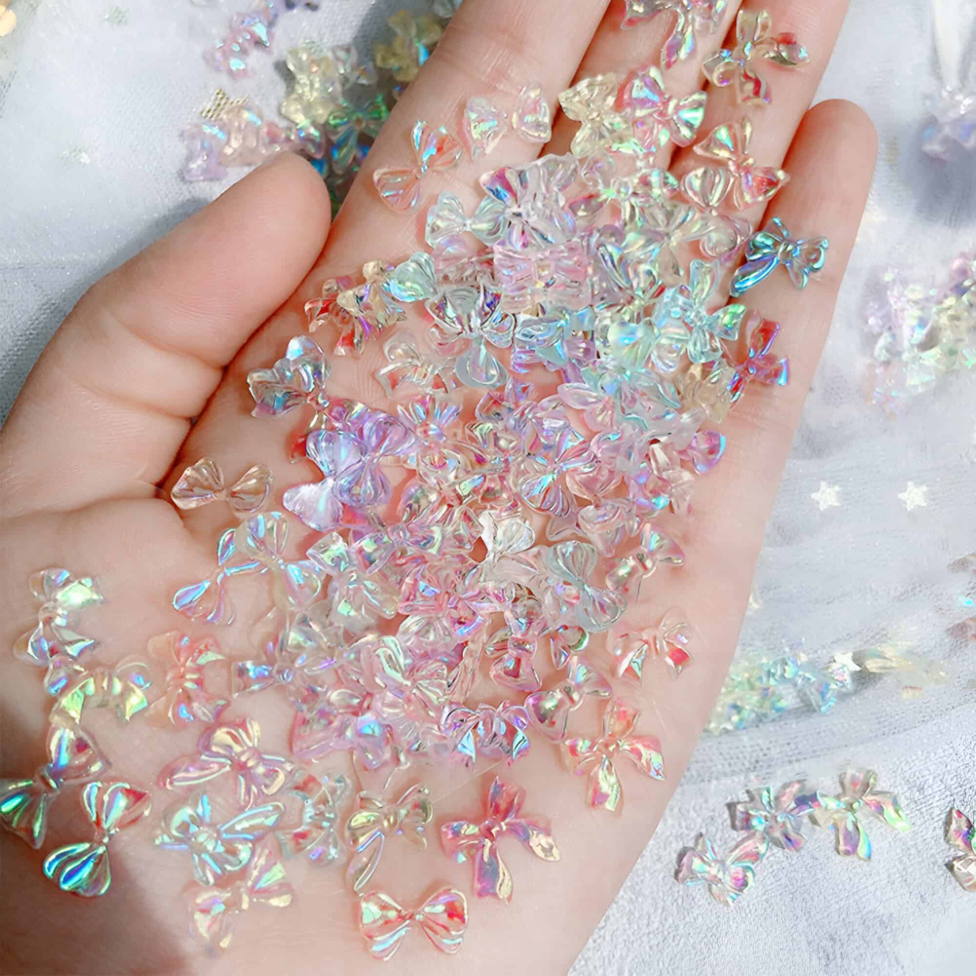 Glittering iridescent bow-shaped nail art embellishments on a hand, acrylic decorations for nails, holographic nail art accessories, Monalisa Medical store quality nail decor, colorful and shiny nail craft supplies.