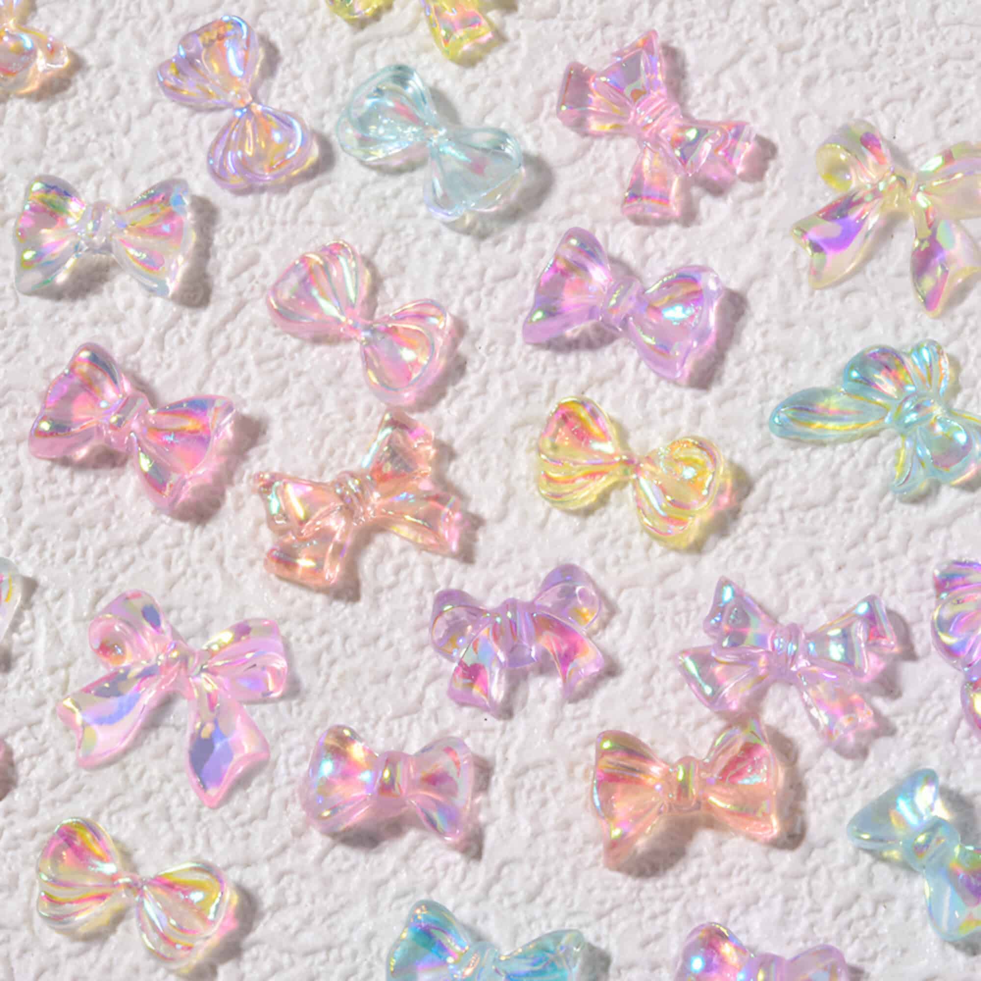 Colorful iridescent bow-shaped charms on textured white background, ideal for nail art, jewelry making, and craft supplies. Trending accessories for personalized, stylish DIY projects.