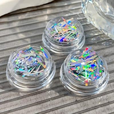 Shimmering holographic nail glitter in small clear containers, ideal for nail art and beauty enhancements. Perfect for adding sparkle and elegance to manicures.