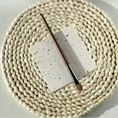Polished makeup brush with wooden handle resting on a textured white stone surface, set against woven round placemat. Elegant cosmetic tools, perfect for beauty and skincare routines.