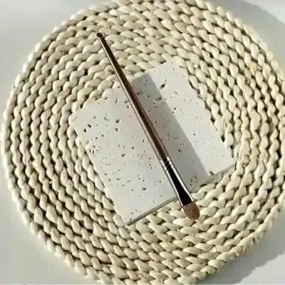 Polished makeup brush with wooden handle resting on a textured white stone surface, set against woven round placemat. Elegant cosmetic tools, perfect for beauty and skincare routines.