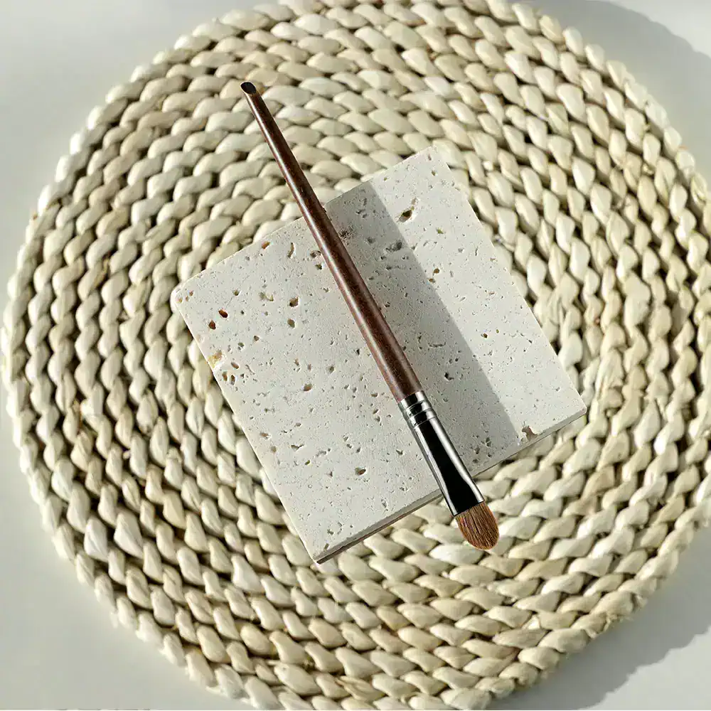 Polished makeup brush with wooden handle resting on a textured white stone surface, set against woven round placemat. Elegant cosmetic tools, perfect for beauty and skincare routines.