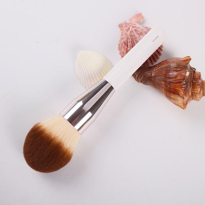 Luxurious makeup brush with a white handle and soft bristles, accompanied by seashells on a white background for skincare or beauty product display.