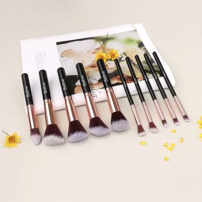 Luxurious makeup brushes set with black handles and rose gold ferrules, arranged on a white surface with colorful flower accents and a Monalisa-inspired magazine background.