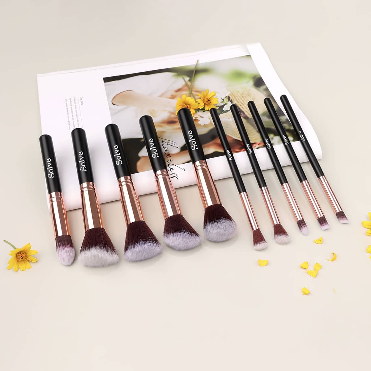 Luxurious makeup brushes set with black handles and rose gold ferrules, arranged on a white surface with colorful flower accents and a Monalisa-inspired magazine background.