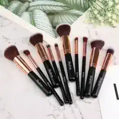 Black and rose gold makeup brushes from Solve laid out on a marble surface with a tropical plant and a botanical print in the background.