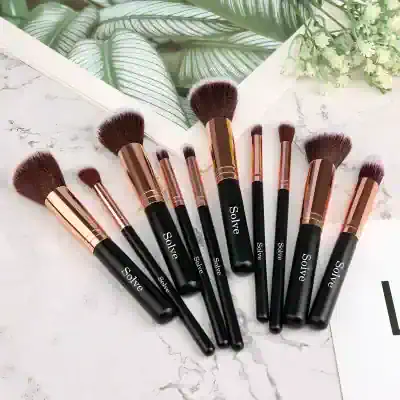Black and rose gold makeup brushes from Solve laid out on a marble surface with a tropical plant and a botanical print in the background.