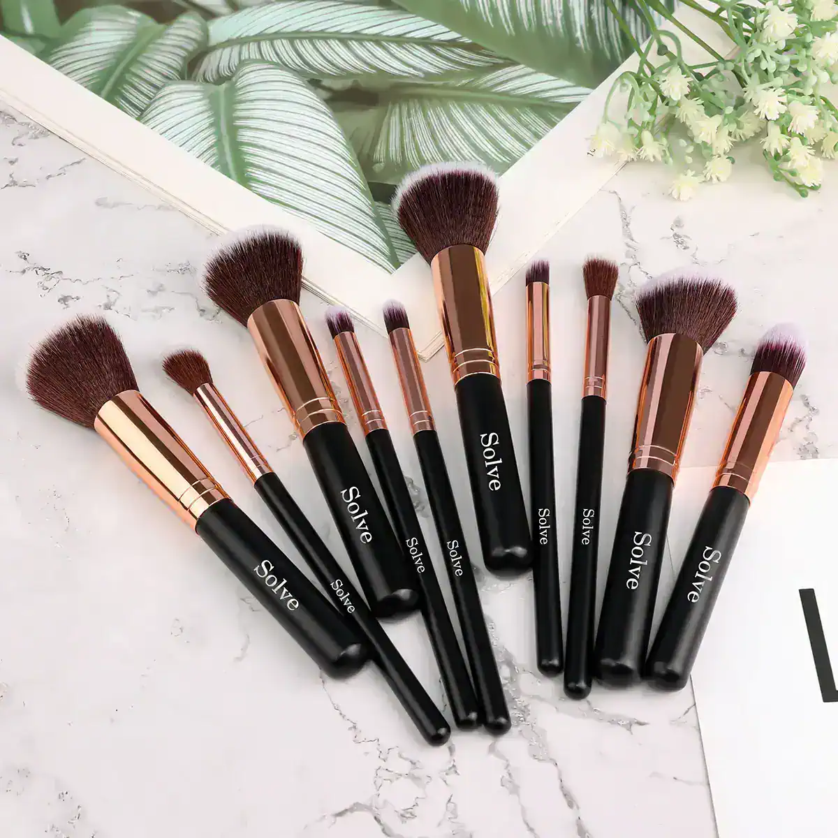 Black and rose gold makeup brushes from Solve laid out on a marble surface with a tropical plant and a botanical print in the background.