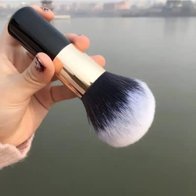 Soft makeup brush with black and white bristles, held outdoors near a body of water, emphasizing beauty and cosmetics.