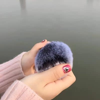 Luxurious faux fur pom-pom hair accessory in soft pastel purple, held by a hands with decorated nails, against a blurred outdoor background. Perfect for stylish hair decor and fashion accessories.
