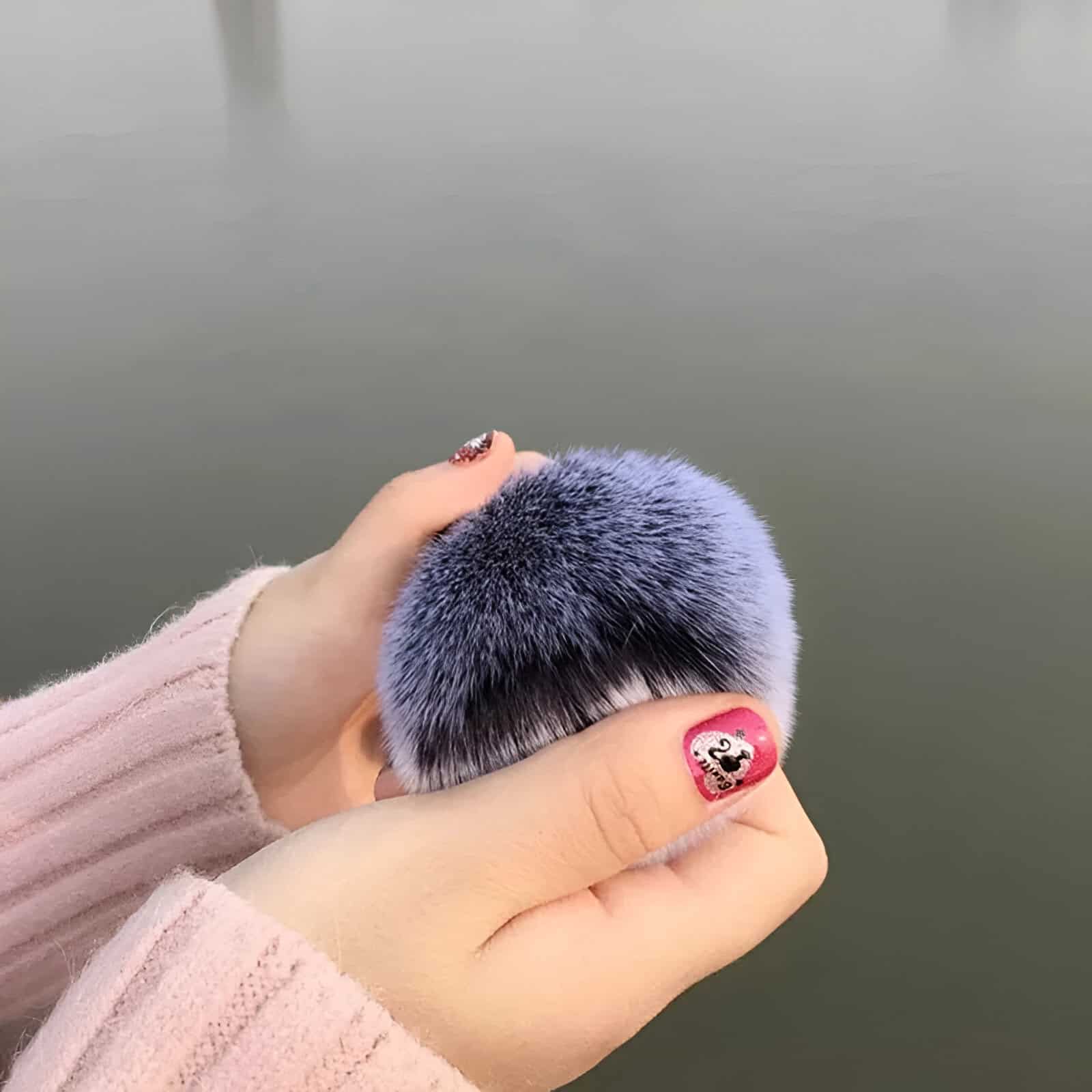 Luxurious faux fur pom-pom hair accessory in soft pastel purple, held by a hands with decorated nails, against a blurred outdoor background. Perfect for stylish hair decor and fashion accessories.