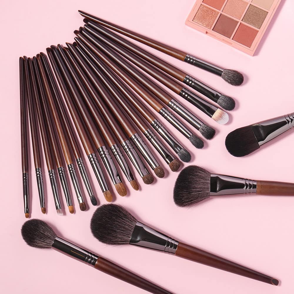 A set of professional makeup brushes with brown handles and black tips, spread out on a pink background alongside a neutral eyeshadow palette, ideal for beauty and cosmetic applications.