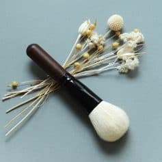 Soft-bristled makeup brush with a wooden handle and white synthetic bristles, surrounded by dried white flowers on a grey background, ideal for cosmetic application.