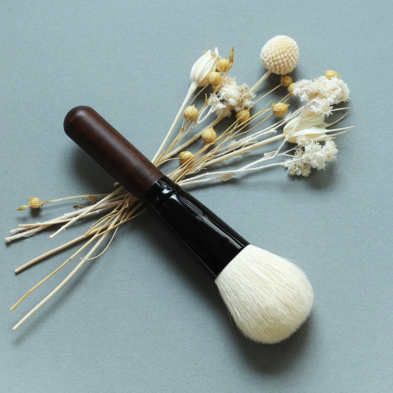 Soft-bristled makeup brush with a wooden handle and white synthetic bristles, surrounded by dried white flowers on a grey background, ideal for cosmetic application.