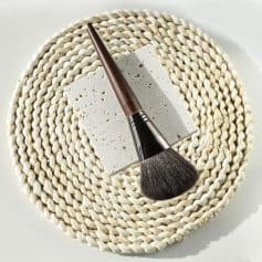 Natural makeup brush with wooden handle on textured stone and woven rattan placemat for skincare and beauty products.