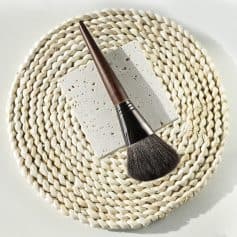 Elegant makeup brush with wooden handle resting on a textured square tile, placed on a woven round mat, ideal for beauty and cosmetic application at Monalisa Store, a trusted destination for skincare and makeup products.