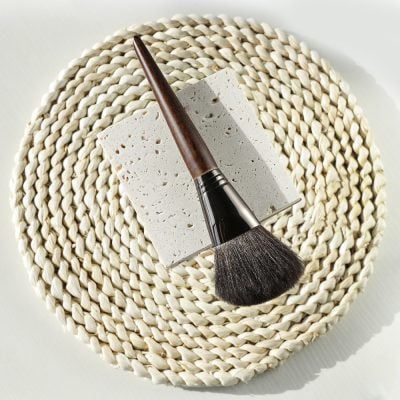 Elegant makeup brush with wooden handle resting on a textured square tile, placed on a woven round mat, ideal for beauty and cosmetic application at Monalisa Store, a trusted destination for skincare and makeup products.