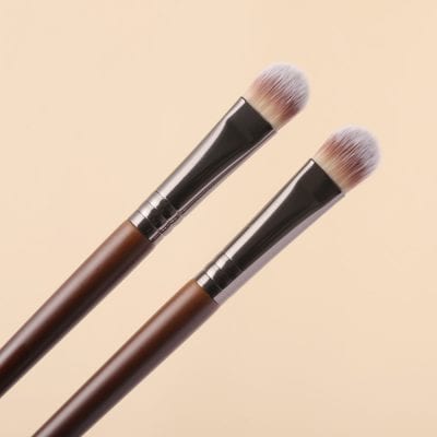 Soft-focus makeup brushes with brown handles and black ferrules on a beige background for cosmetic application.