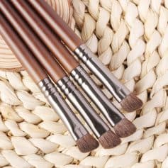 Professional makeup brushes with wooden handles and synthetic bristles on woven natural background, ideal for beauty and cosmetic application.