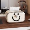 Smiley-face tissue box holder with tissue paper, next to headphones and a camera on a wooden desk, ideal for optometry or eyewear store decor.