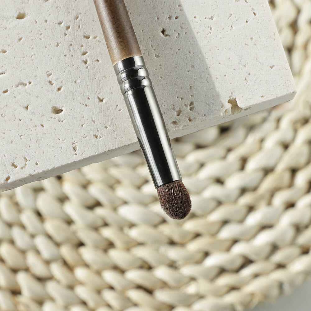 Soft makeup brush with wooden handle and metal ferrule resting on a textured beige surface at Monalisa Store.