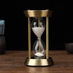 Shaped hourglass timer with gold frame and clear glass, filled with white sand, on wooden surface against black background, symbolizing time management and precision.