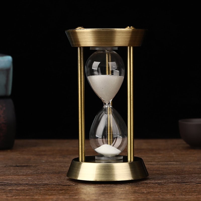 Shaped hourglass timer with gold frame and clear glass, filled with white sand, on wooden surface against black background, symbolizing time management and precision.